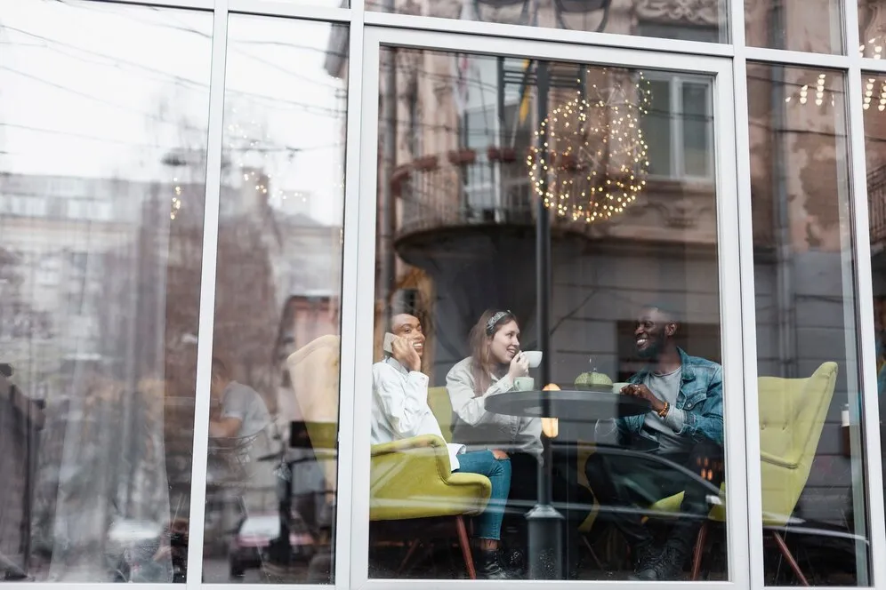 Friends sitting near a window in a coffee shop, representing the importance of strengthening the business's brand with 2025 commercial windows and doors trends.